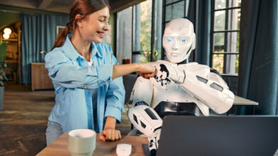 Darstellung Zusammenarbeit Mensch und KI: Frau gibt Roboter der am Laptop im Büro sitzt ein Fist Bump